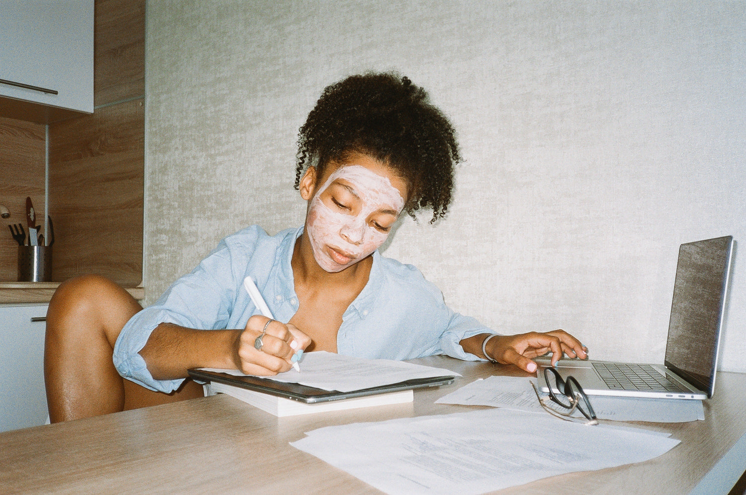 Picture of woman working from home in front of a laptop and making notes on a notepad while wearing her hair up and face mask on.