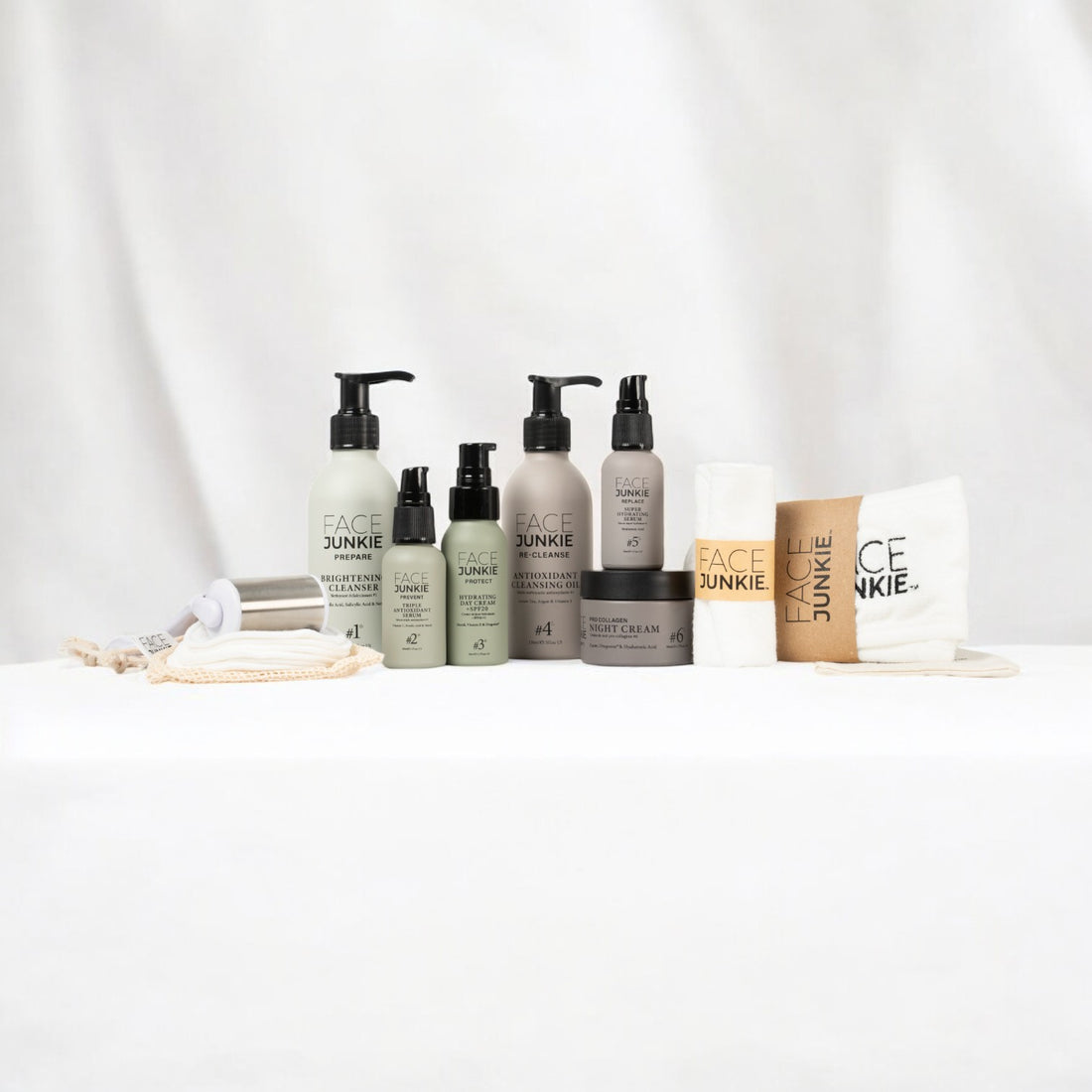 Ultimate Hero Collection Skincare Bundle by Face Junkie displayed on a white surface with a soft fabric background. The set includes various skincare products such as brightening cleanser, antioxidant cleansing oil, night cream and facial serum, all in minimalist packaging with earthy tones. Accompanying accessories include reusable face cloths, a face roller, and reusable bamboo pads. The aesthetic is clean and eco-friendly, appealing to natural skincare enthusiasts.