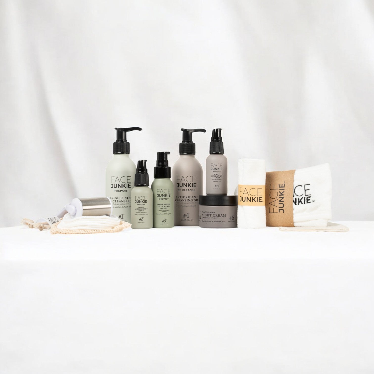 Ultimate Hero Collection Skincare Bundle by Face Junkie displayed on a white surface with a soft fabric background. The set includes various skincare products such as brightening cleanser, antioxidant cleansing oil, night cream and facial serum, all in minimalist packaging with earthy tones. Accompanying accessories include reusable face cloths, a face roller, and reusable bamboo pads. The aesthetic is clean and eco-friendly, appealing to natural skincare enthusiasts.