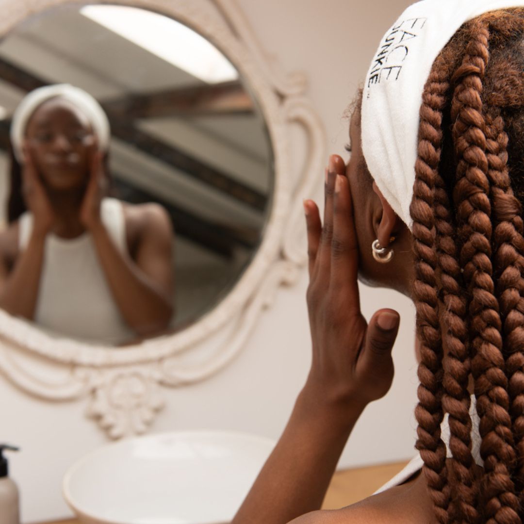Close-up of a woman applying skincare in front of a mirror, wearing a white Face Junkie headband. Her skin looks radiant as she gently massages a product into her face. The image highlights the ritual of self-care and skincare with Face Junkie’s high-performance products, promoting healthy, glowing skin. The aesthetic includes a minimalistic bathroom setting with a white sink and skincare products visible on the counter.