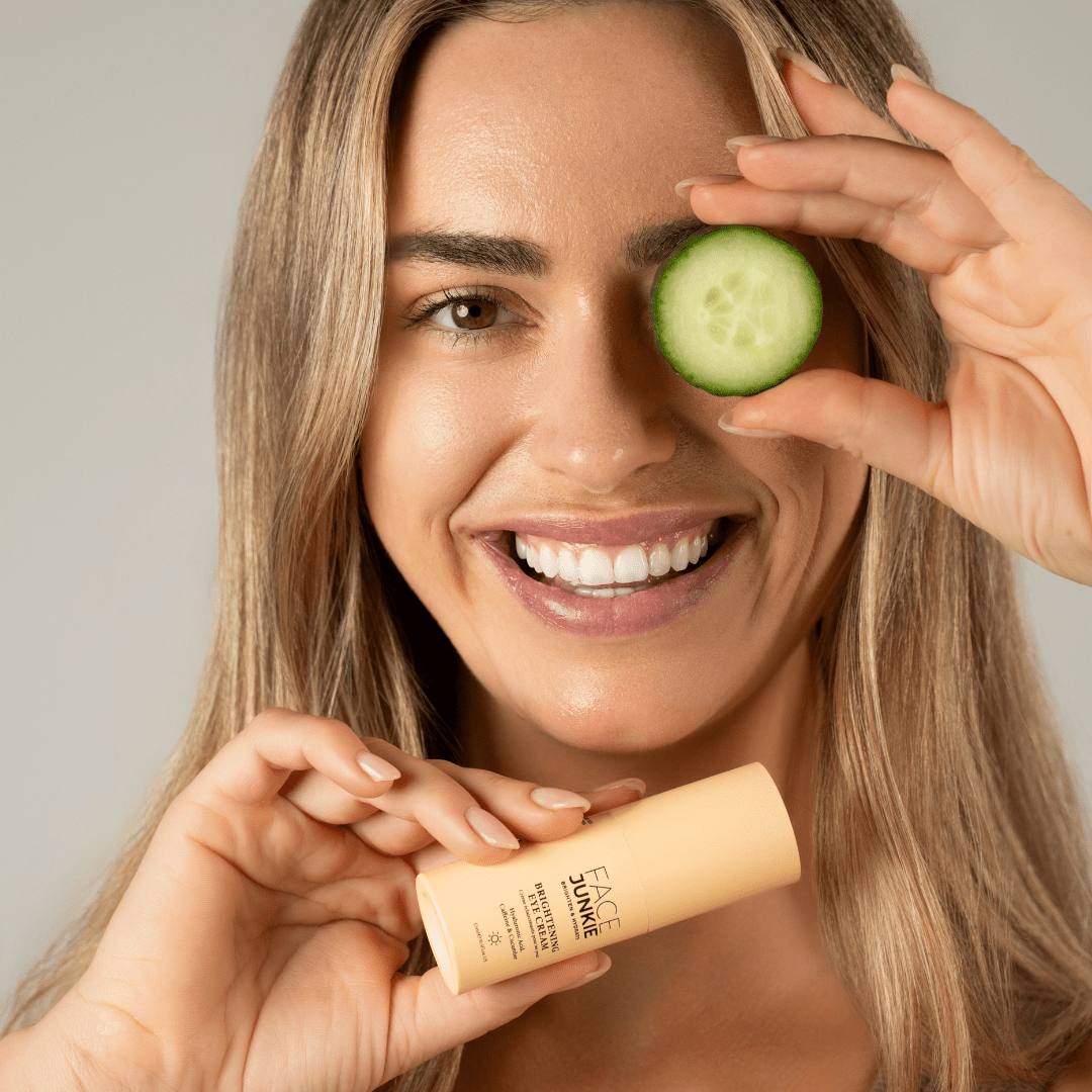 A smiling woman holds a cucumber slice over one eye while holding a bottle of "FACE JUNKIE Brightening Eye Cream" in her other hand. The woman has long, light brown hair and is showcasing a radiant complexion. The product is in a light peach-colored bottle, and the overall vibe of the image is fresh and playful, emphasizing skincare and eye care routines.