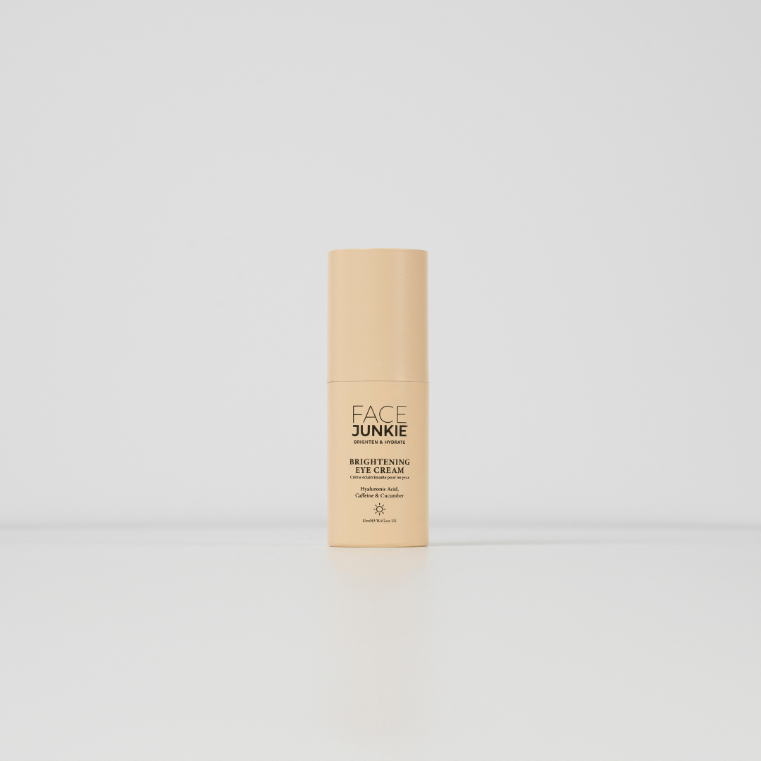 A minimalistic product shot of "FACE JUNKIE Brightening Eye Cream" in a light peach-colored bottle. The bottle stands on a clean white surface with a plain white background, emphasizing the product&