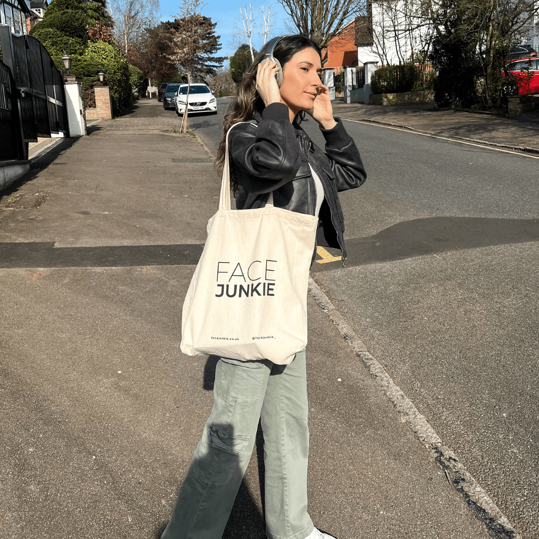 A woman walks down a sunny suburban street while wearing headphones and carrying a beige FACE JUNKIE tote bag. She is dressed casually in light green pants and a black leather jacket, enjoying the outdoors. The tote bag prominently features the FACE JUNKIE logo, along with the brand&
