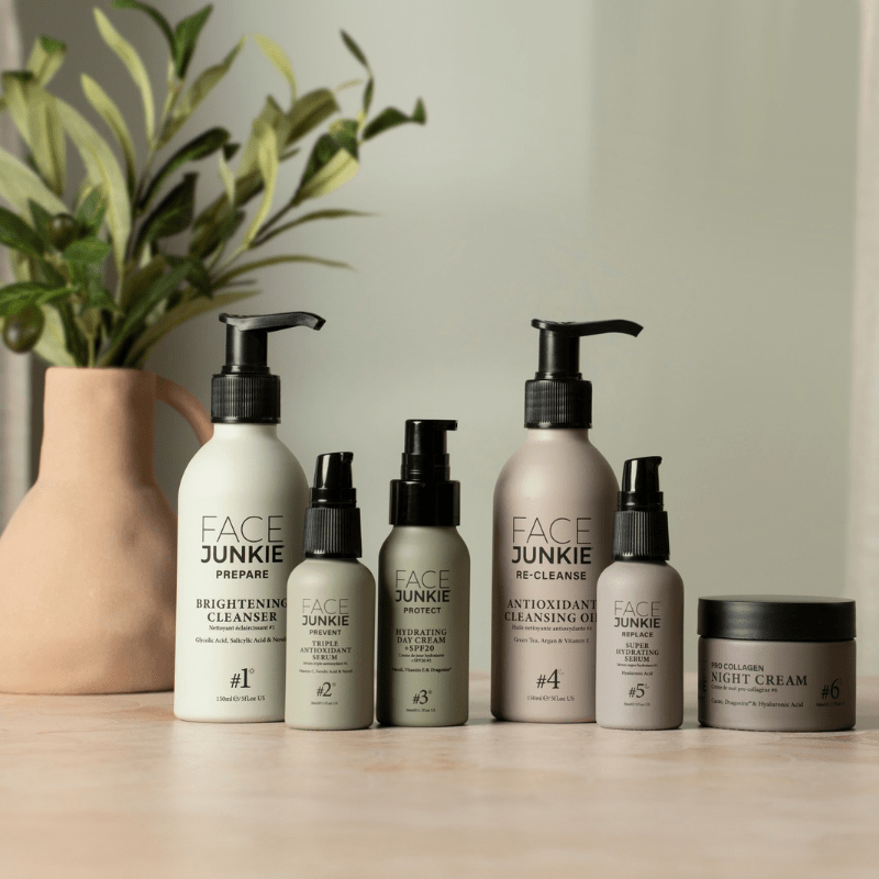 A collection of Face Junkie skincare products arranged on a light wooden surface. The lineup includes cleansers, serums, a day cream with SPF, a night cream, and an antioxidant cleansing oil. The sleek packaging in neutral shades of white and gray gives a minimalist, premium feel. In the background, a muted peach vase with green foliage adds a natural touch, complementing the skincare products&