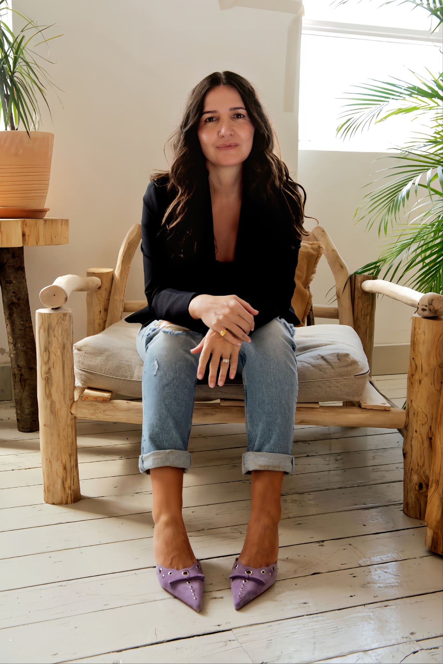 Portrait of a woman sitting on a wooden chair in a stylish, minimalist space. She wears a black blazer, ripped jeans, and purple pointed heels, showcasing her casual yet sophisticated look. The background features natural light and greenery, emphasizing a relaxed, contemporary vibe. The woman’s confident and calm expression complements the holistic lifestyle promoted by Face Junkie’s skincare products.