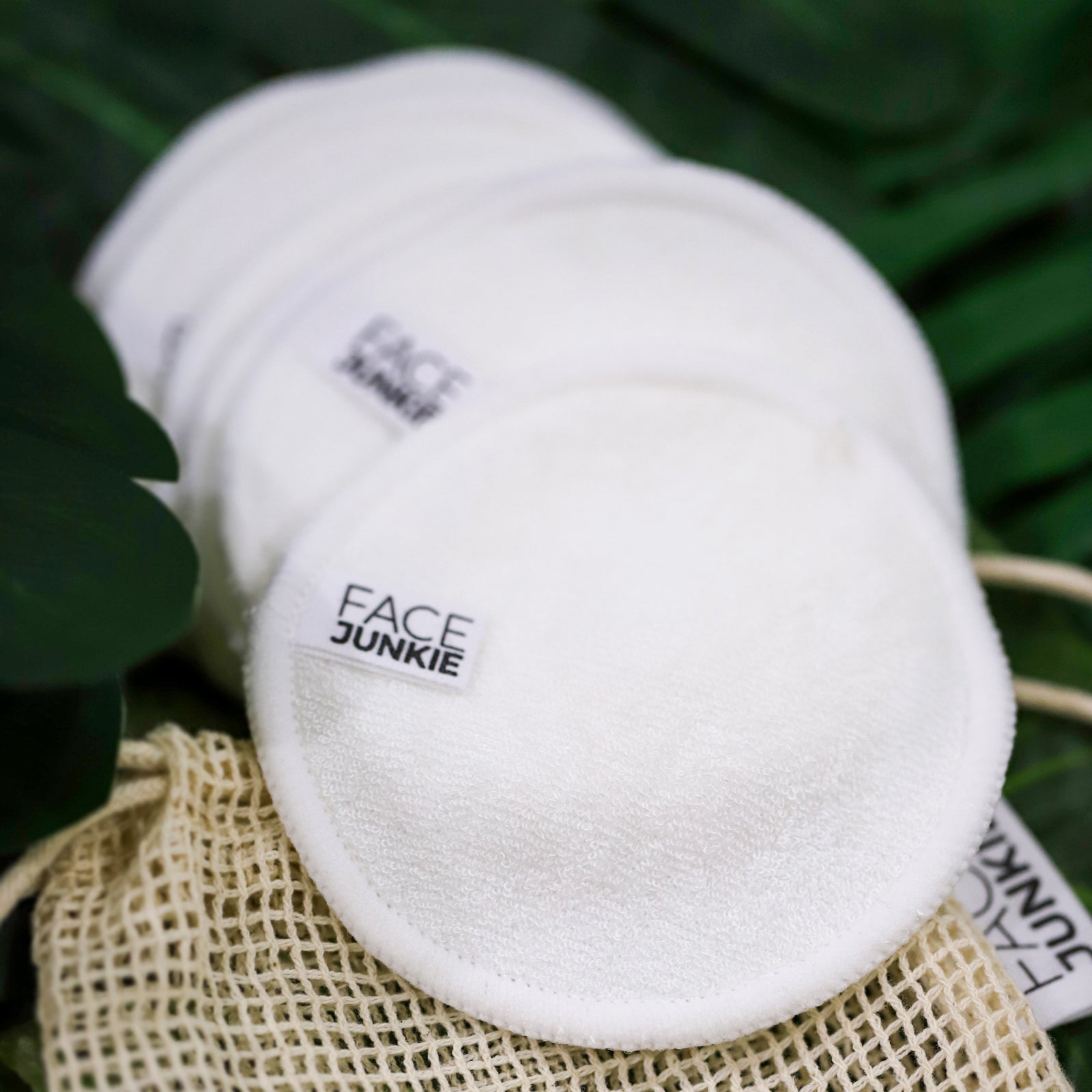 A close-up of a set of white reusable FACE JUNKIE bamboo cleansing pads stacked together on a natural mesh bag. The soft pads feature the brand&