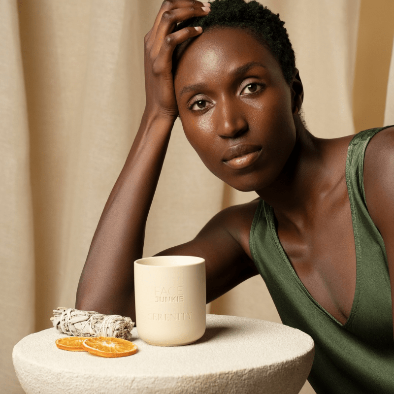 A woman with radiant skin and short natural hair, wearing a green sleeveless top, poses thoughtfully while resting her head on her hand. In front of her on a textured white table is a Face Junkie "SERENITY" candle, along with dried sage and slices of dried orange. The neutral-toned background enhances the peaceful, self-care ambiance of the scene, highlighting the calming and luxurious aesthetic of the Face Junkie brand.