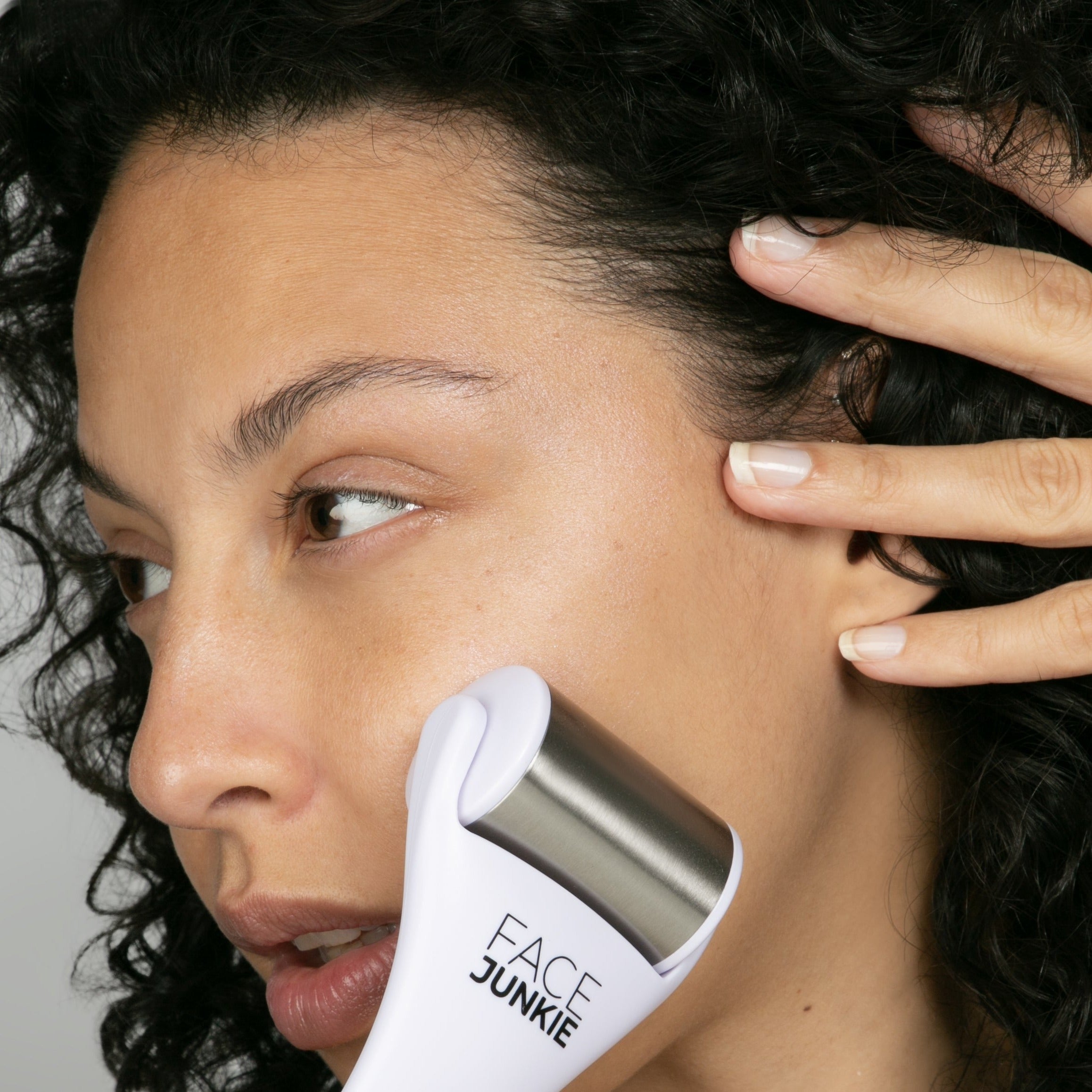 A close-up of a woman with curly hair using a Face Junkie-branded facial roller on her cheek. The stainless steel roller is gently applied to her skin, promoting circulation and skin rejuvenation. Her hand is positioned to hold her hair back, highlighting her natural complexion and the smooth, calming effect of the skincare tool. The image conveys a sense of relaxation and self-care as part of a skincare routine.
