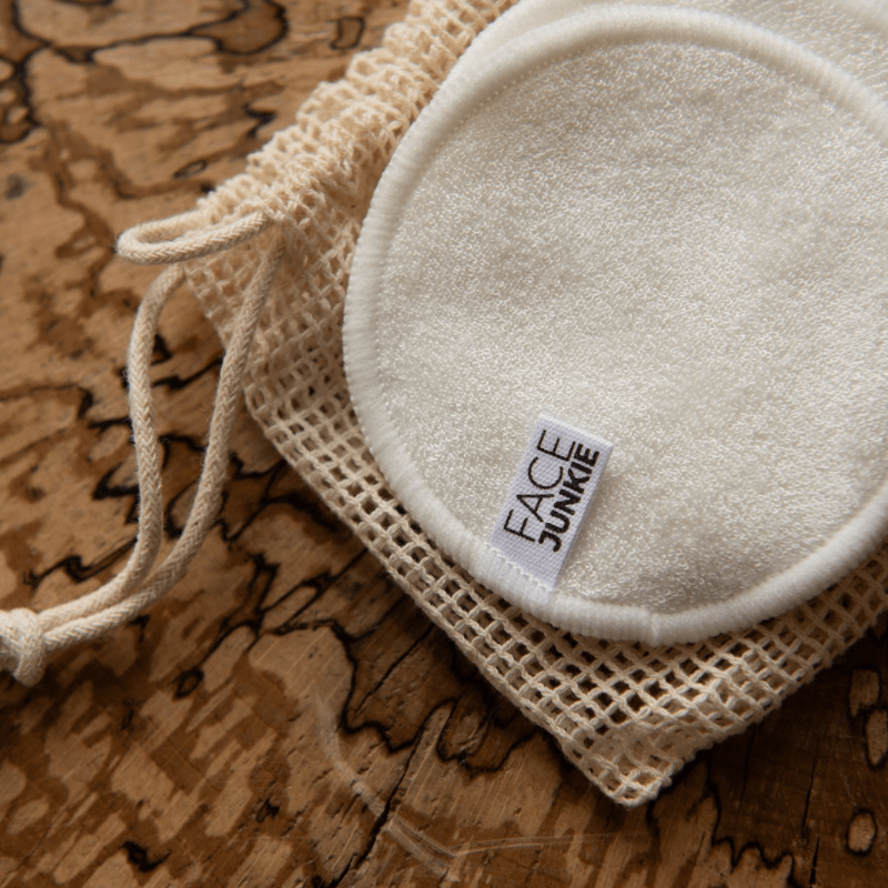 A set of reusable, soft white face pads from the brand Face Junkie, displayed on a rustic wooden surface. The pads are accompanied by a mesh drawstring bag, giving a natural and eco-friendly vibe. The Face Junkie logo is visible on one of the pads, highlighting the brand&