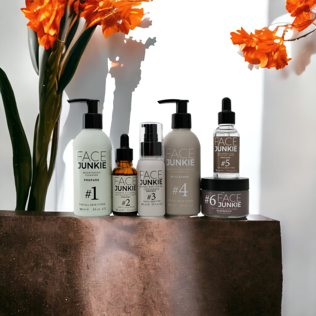 Face Junkie skincare collection beautifully displayed with vibrant orange flowers in the background. The lineup includes the Brightening Cleanser (#1), Hydrating Vitamin C Serum (#2), Hydrating Day Cream with SPF 20 (#3), Antioxidant Cleansing Oil (#4), Repairing Peptide Serum (#5), and Pro Collagen Night Cream (#6). Each product is designed for all skin types and features clean, high-performance ingredients for a holistic skincare routine promoting healthy, radiant skin.