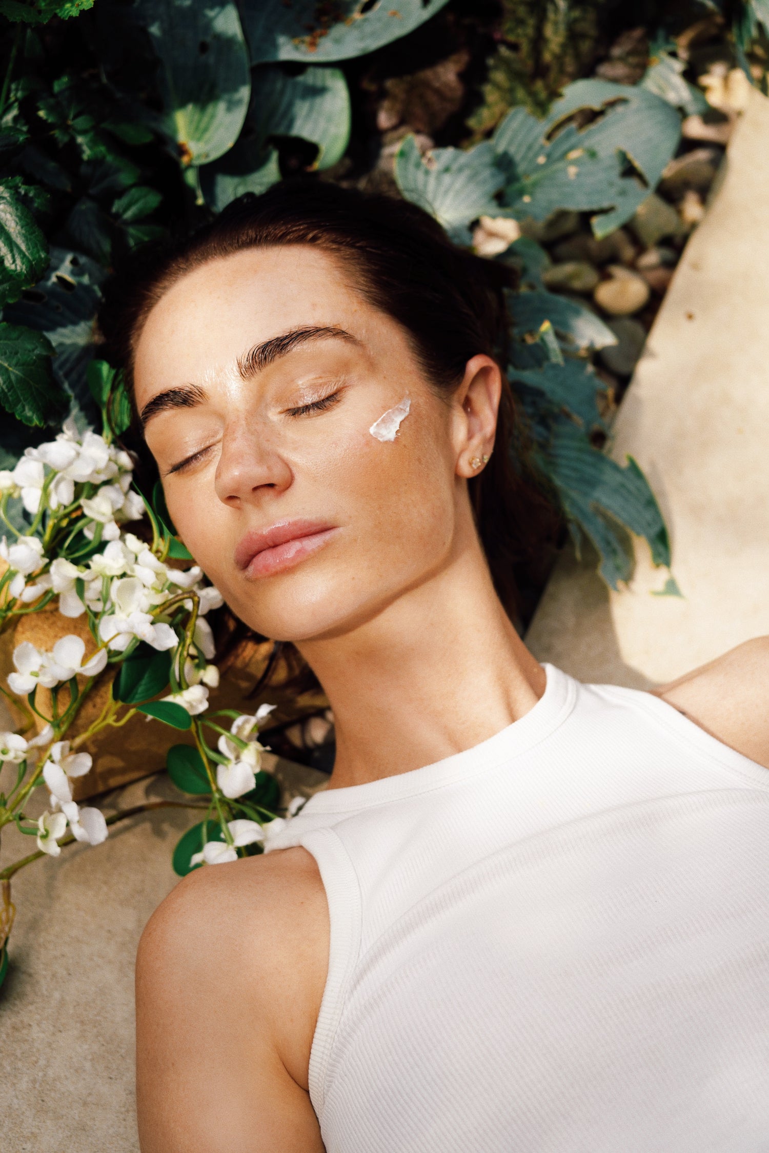 A woman with glowing skin laying down on a bed of plants and flowers, applying skincare product to her cheek.