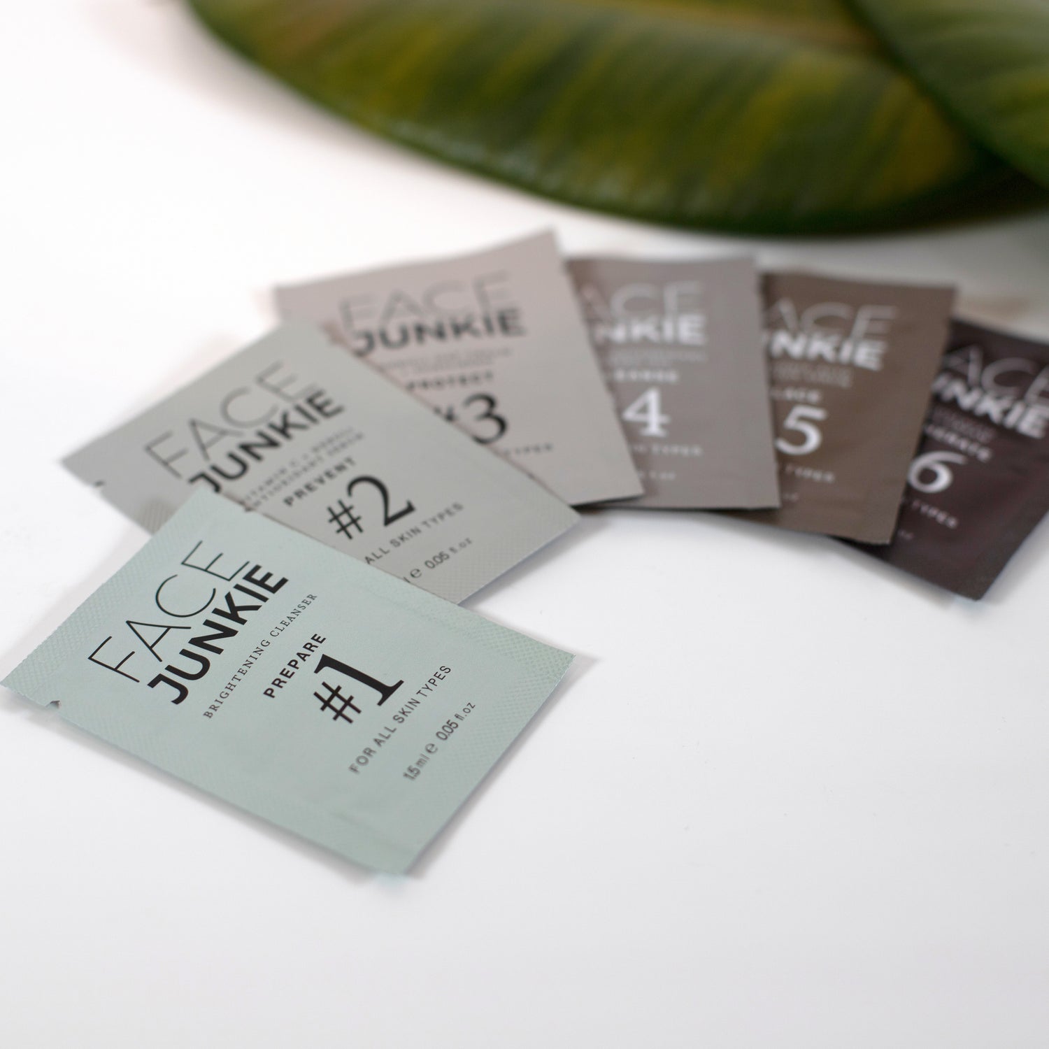 Picture of all six of the Face Junkie Samples for the Hero Collection fanned out on a white table with green earthy leaves in view.