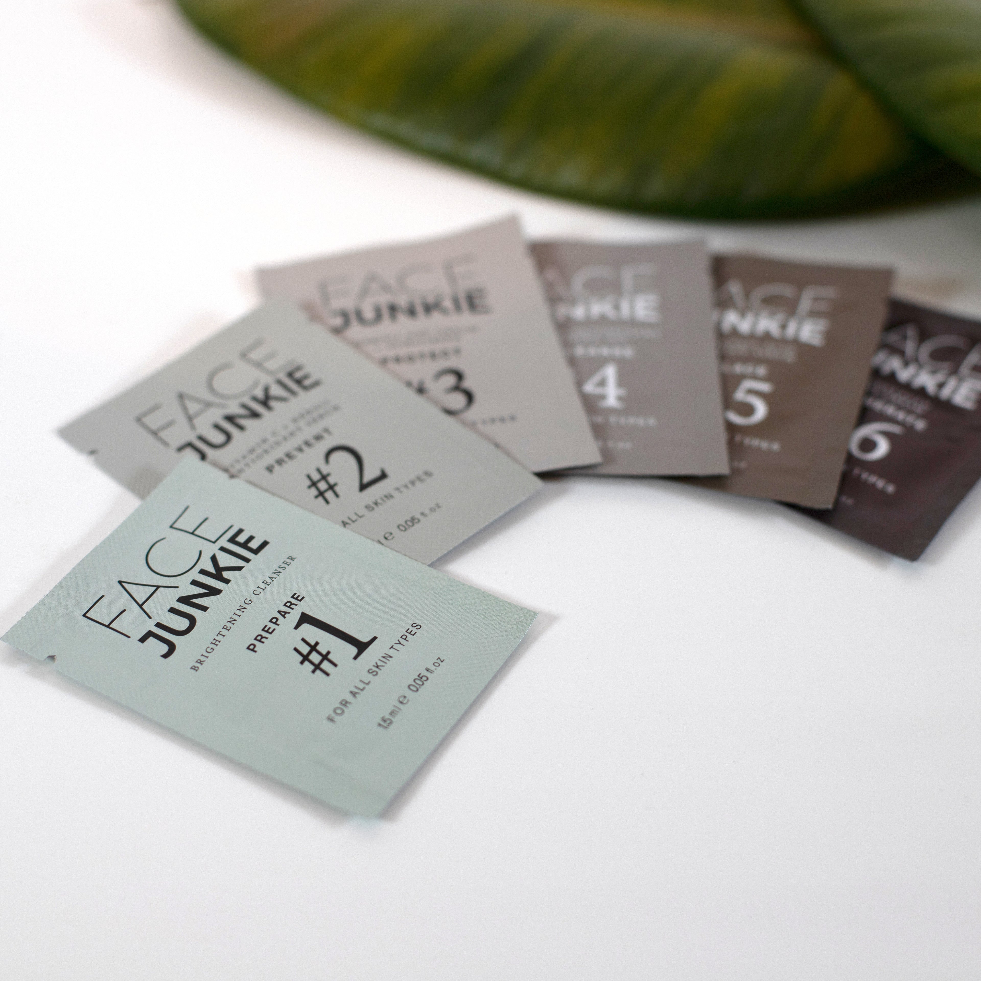 Picture of all six of the Face Junkie Samples for the Hero Collection fanned out on a white table with green earthy leaves in view.