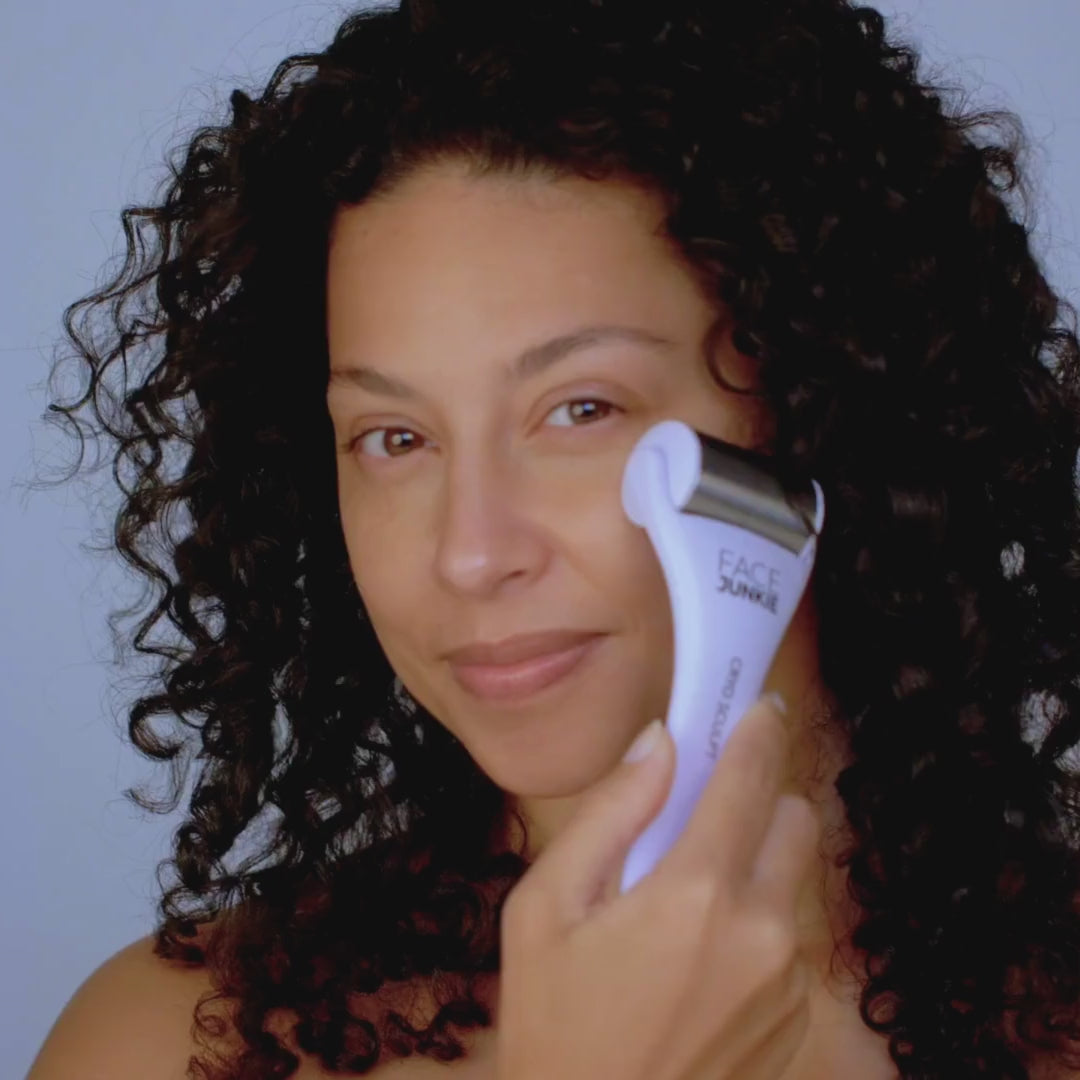 A woman with curly dark hair uses a FACE JUNKIE facial roller on her cheek while smiling gently at the camera. The woman has a natural, radiant look and is in the process of performing a skincare routine. The facial roller is white with a metallic roller head and the brand&
